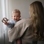 Bébé souriant tenant un anneau de dentition bretzel Mushie brun, porté par sa maman
