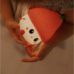 Un enfant tient une jolie fraise veilleuse en forme de personnage souriant avec un chapeau de fraise, avec de grands yeux et des joues roses - cette veilleuse USB rechargeable est de la marque Marys.