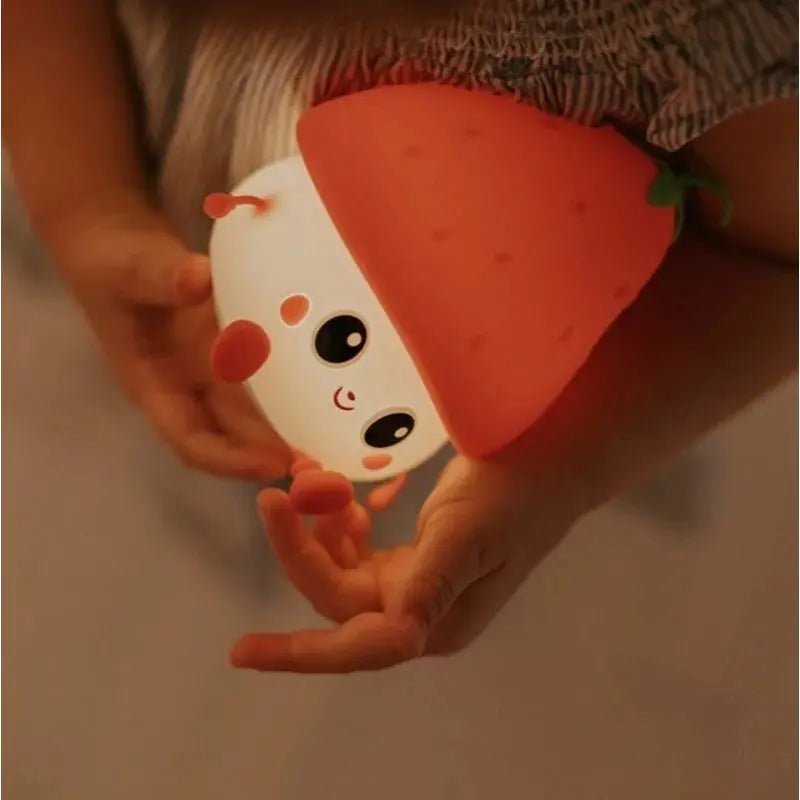 Un enfant tient une jolie fraise veilleuse en forme de personnage souriant avec un chapeau de fraise, avec de grands yeux et des joues roses - cette veilleuse USB rechargeable est de la marque Marys.