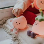 La main d'un enfant tient une veilleuse pêche, une veilleuse en forme de pêche avec un visage souriant. Cette veilleuse USB rechargeable Mary's se trouve sur un tapis texturé à côté d'une autre lampe en forme de fruit.