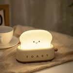 Une veilleuse en forme de tranche de pain avec un visage souriant, étiquetée "TOAST", se trouve sur une table à côté d'une tasse et d'une soucoupe.