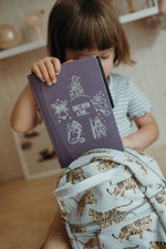 Un jeune enfant en chemise rayée place un livre d'histoires violet intitulé Once Upon A Time et un dessin de tablette Kidywolf dans un sac à dos blanc avec un motif imprimé tigre. Le visage de l'enfant est partiellement obscurci par le livre.